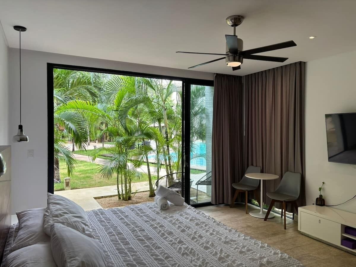 A modern bedroom with a large bed, a ceiling fan, a TV, and a view of a pool and palm trees.