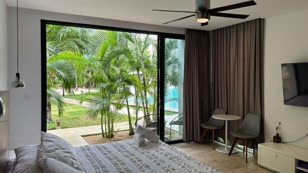 Un dormitorio moderno con una cama grande, un ventilador de techo, un televisor y vistas a una piscina y palmeras.