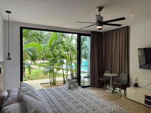 A modern bedroom with a large bed, a ceiling fan, a TV, and a view of a pool and palm trees. - Casita Oleaje Luxury Cultural Sanctuary & Spa (Tulum)