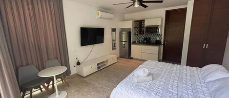 A hotel room with a bed, a desk, a chair, a television, an air conditioning unit, and a kitchenette.