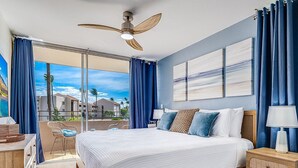 A bedroom with a large bed, a ceiling fan, a nightstand, and a view of the outdoors. - Beachfront 4BR, 2 Condos for 12 in Maalaea! (Wailuku)