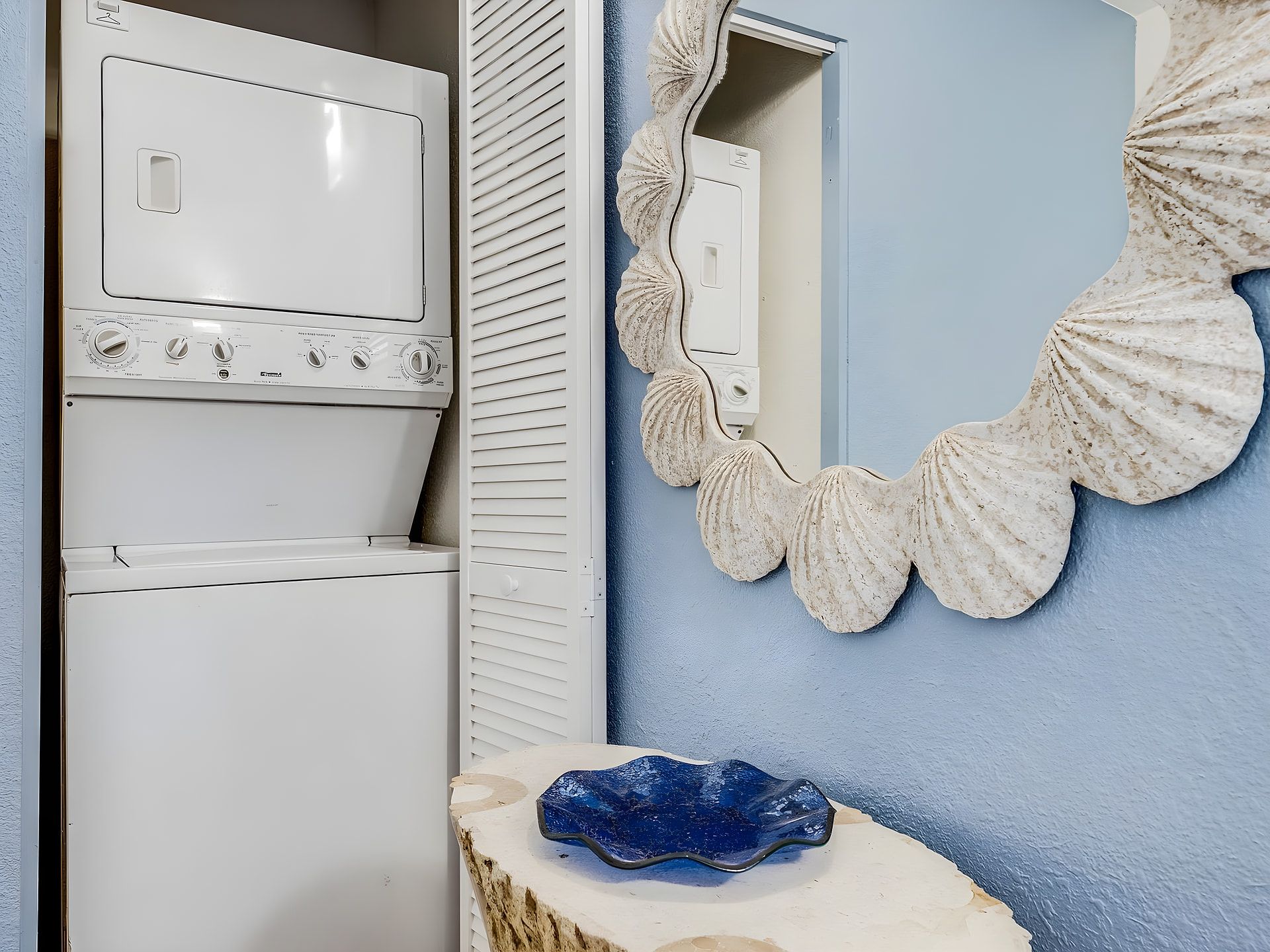 A laundry room with stacked washing machines, a mirror with a shell design, and a blue decorative piece on a white surface.