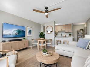 A modern living room with a flat-screen TV, a ceiling fan, a dining area with a round table, and a kitchen with wooden cabinets. - Maalaea, Island Sands, Spacious 4 BR, Beachfront, Sleeps 12 (Wailuku)