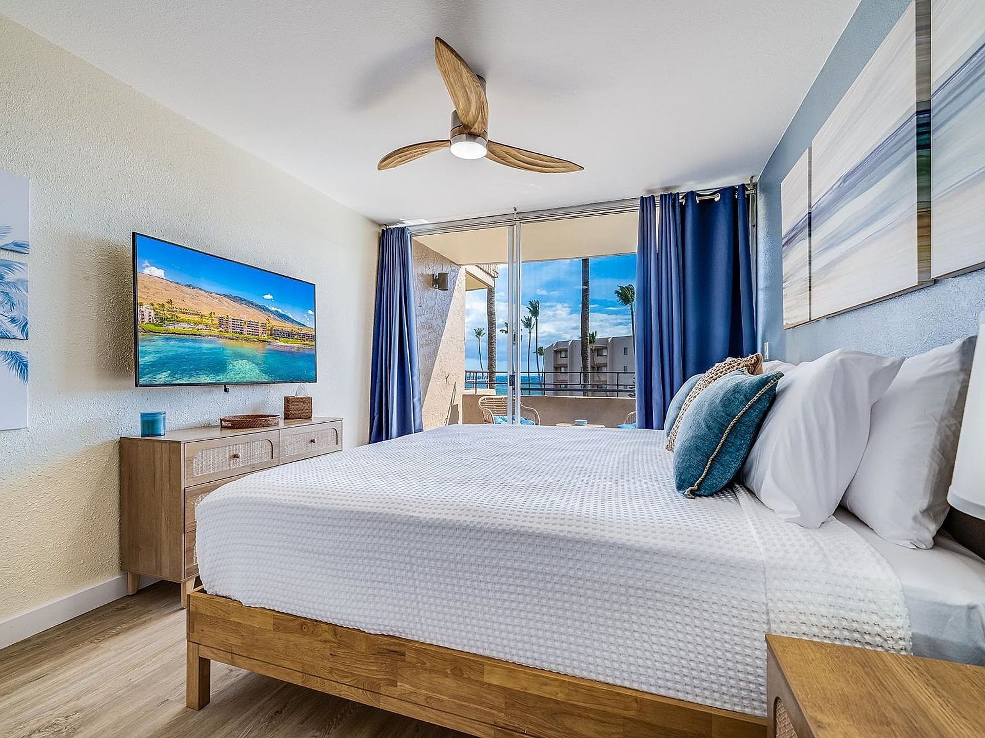 A bedroom with a bed, wooden headboard, a ceiling fan, a TV, and a view of the outdoors.