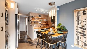 A modern kitchen with a dining area, featuring a wooden table, chairs, and a variety of kitchen appliances. - Beachfront 4BR, 2 Condos for 12 in Maalaea! (Wailuku)