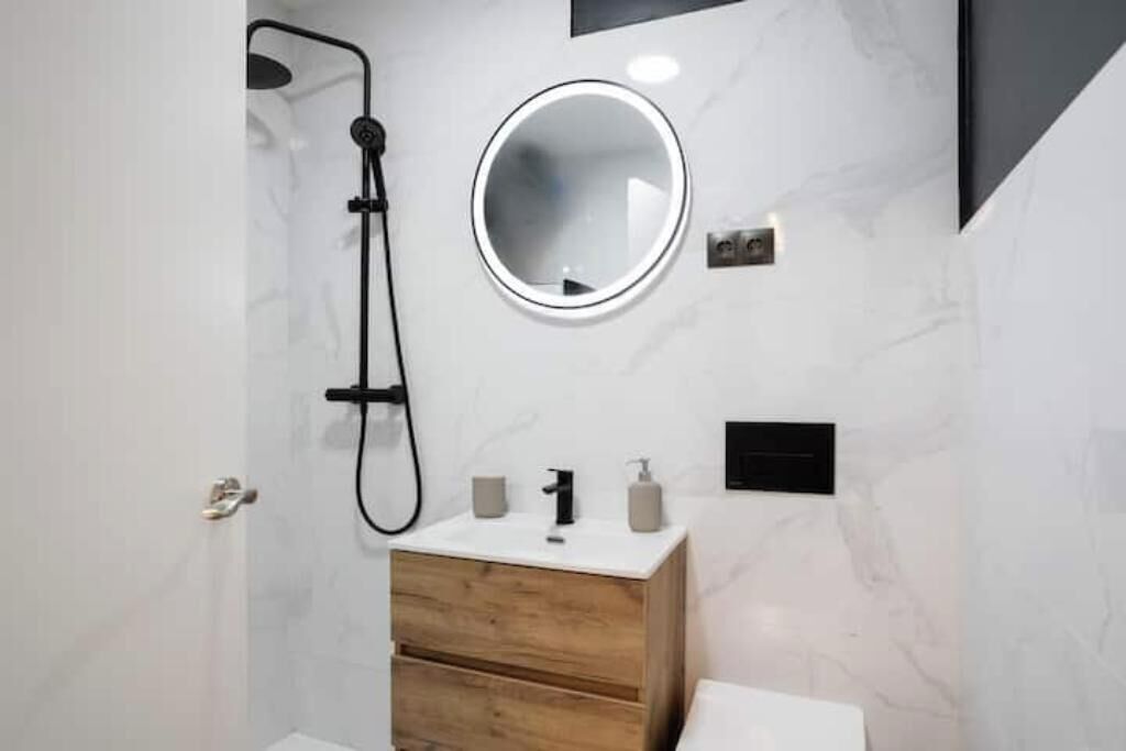 A modern bathroom with a shower, a round mirror, a wooden vanity, and a toilet.