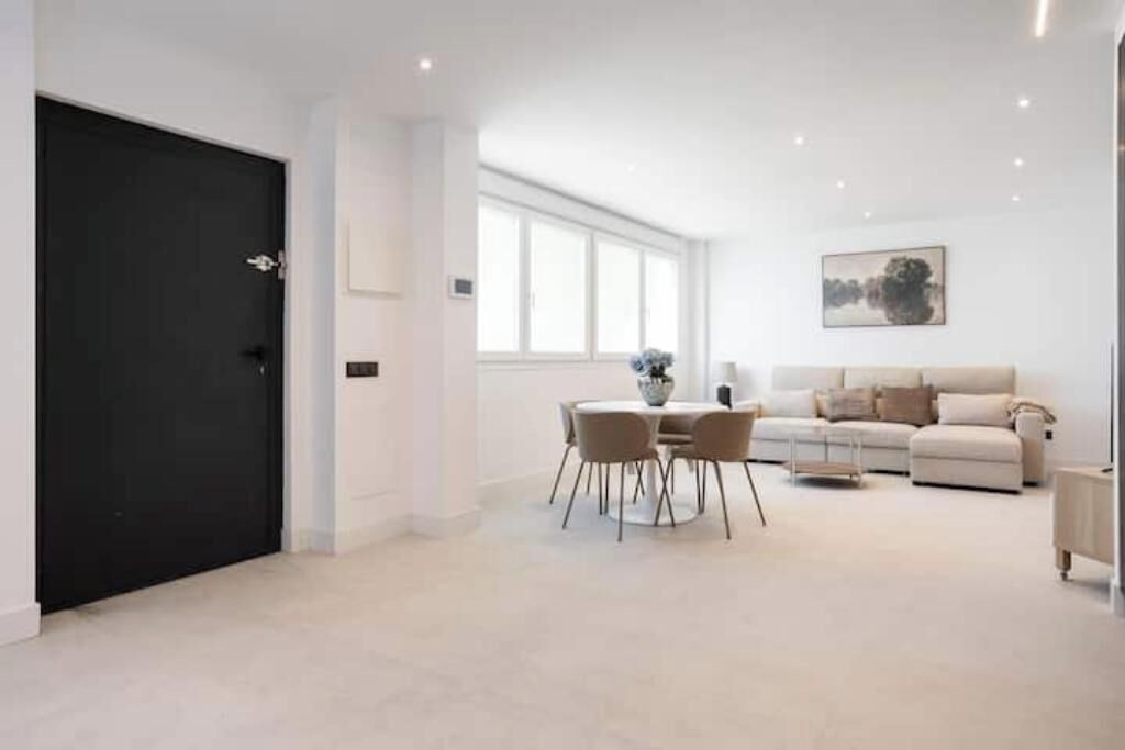 A modern living room with a black door, a round dining table with chairs, a beige sofa, and a large window.