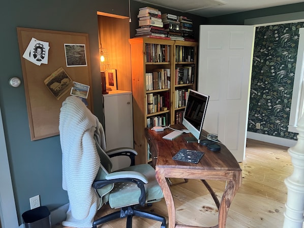 A home office with a desk, chair, computer, bookshelf, and a corkboard with photos and notes.