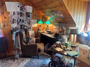 A cozy room with a wooden ceiling, a desk with various items, a chair, and a patterned rug.