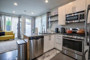 A modern kitchen with stainless steel appliances, a dishwasher, and a dining area with a yellow sofa. - Private Rooftop w Grill, AC + Free Parking (Seattle)