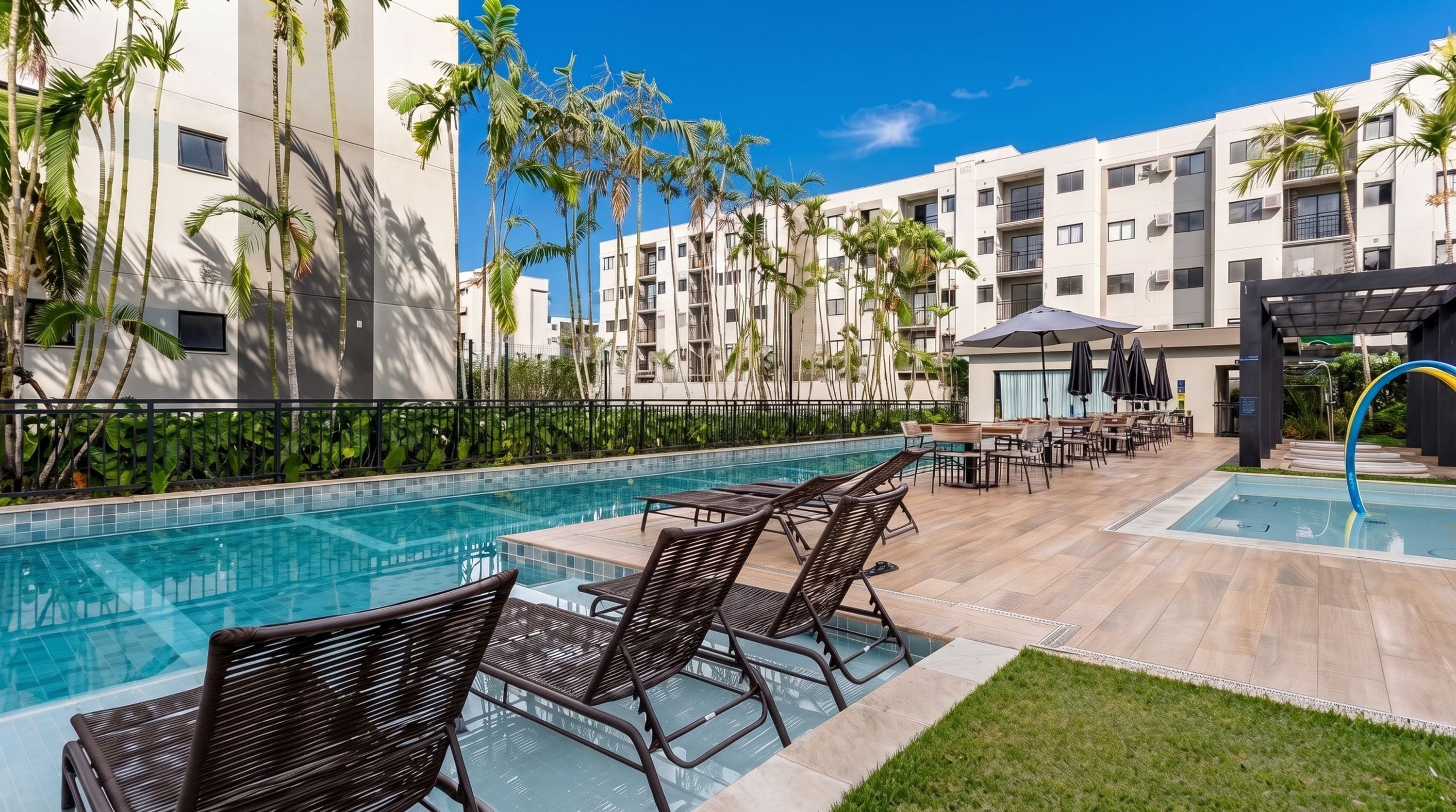A pool area with lounge chairs and umbrellas, surrounded by palm trees and a modern building.