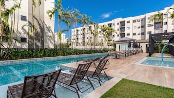 Uma área de piscina com espreguiçadeiras e guarda-sóis, cercada por palmeiras e um edifício moderno.