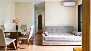 A modern living room with a glass dining table, two chairs, a sofa, and an air conditioning unit mounted on the wall. - Apto c Piscina e Varanda Perto do Parque Ol Mpico (Rio de Janeiro)