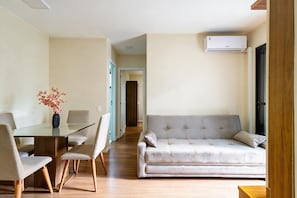 A modern living room with a glass dining table, two chairs, a sofa, and an air conditioning unit mounted on the wall. - Apto c Piscina e Varanda Perto do Parque Ol Mpico (Rio de Janeiro)