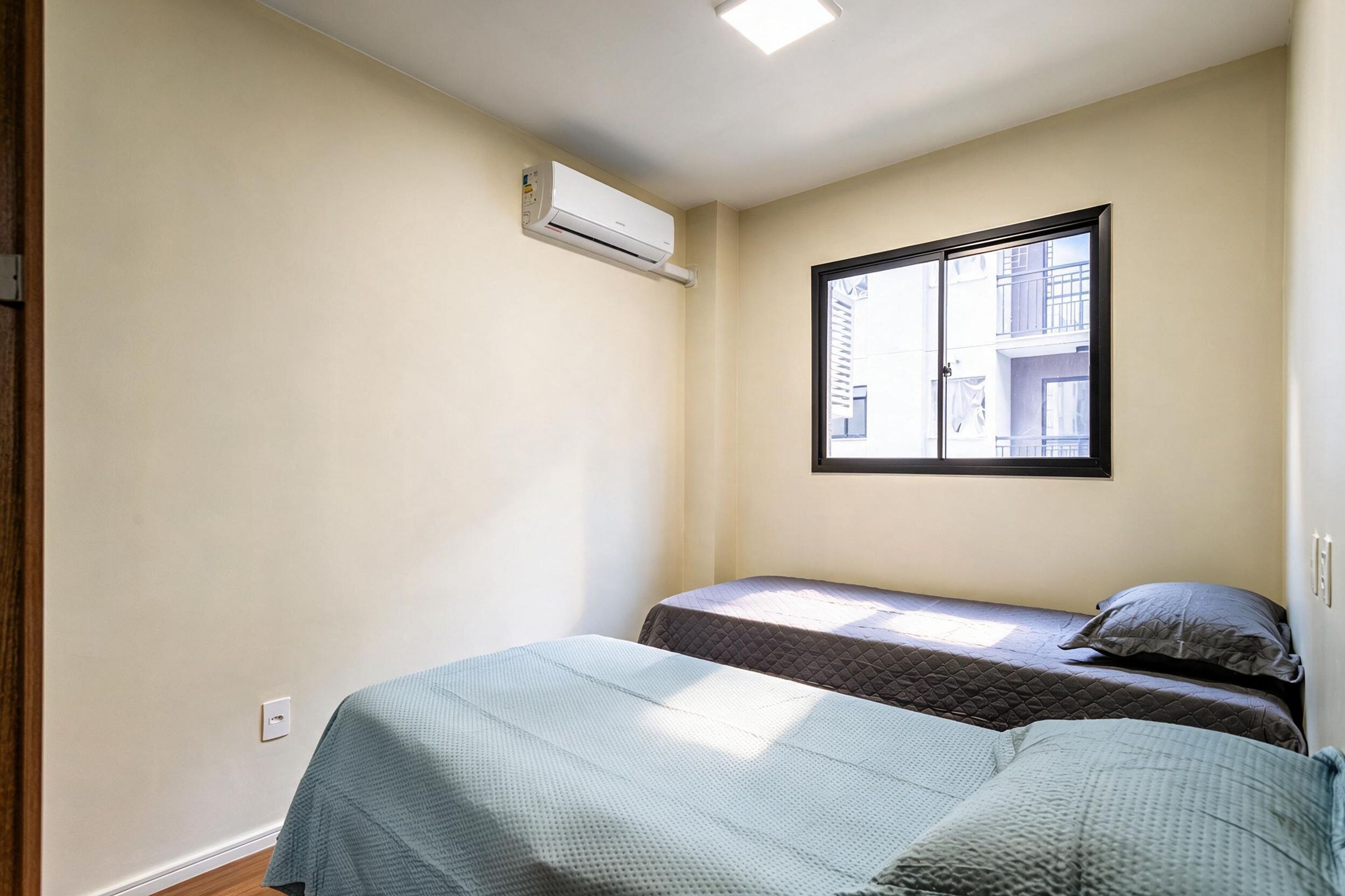 Um pequeno apartamento de um único quarto, com duas camas, uma janela e um aparelho de ar condicionado.