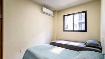 Um pequeno apartamento de um único quarto, com duas camas, uma janela e um aparelho de ar condicionado.