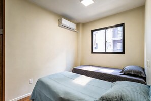 Um pequeno apartamento de um único quarto, com duas camas, uma janela e um aparelho de ar condicionado.