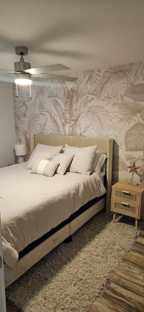 Ein Schlafzimmer mit einem Bett, einem Nachttisch, einer Lampe, einem Deckenventilator und einer Wand mit einem tropischen Blattmuster.