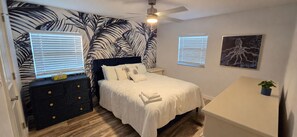 Ein Schlafzimmer mit einem Bett, einem Nachttisch, einer Kommode, einem Deckenventilator und einer Wand mit Blättermotiv.