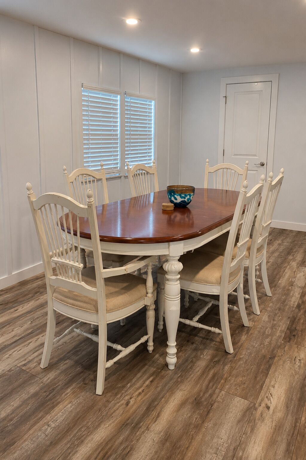 A dining room with a wooden table and white chairs, a bowl on the table, and a door in the background.