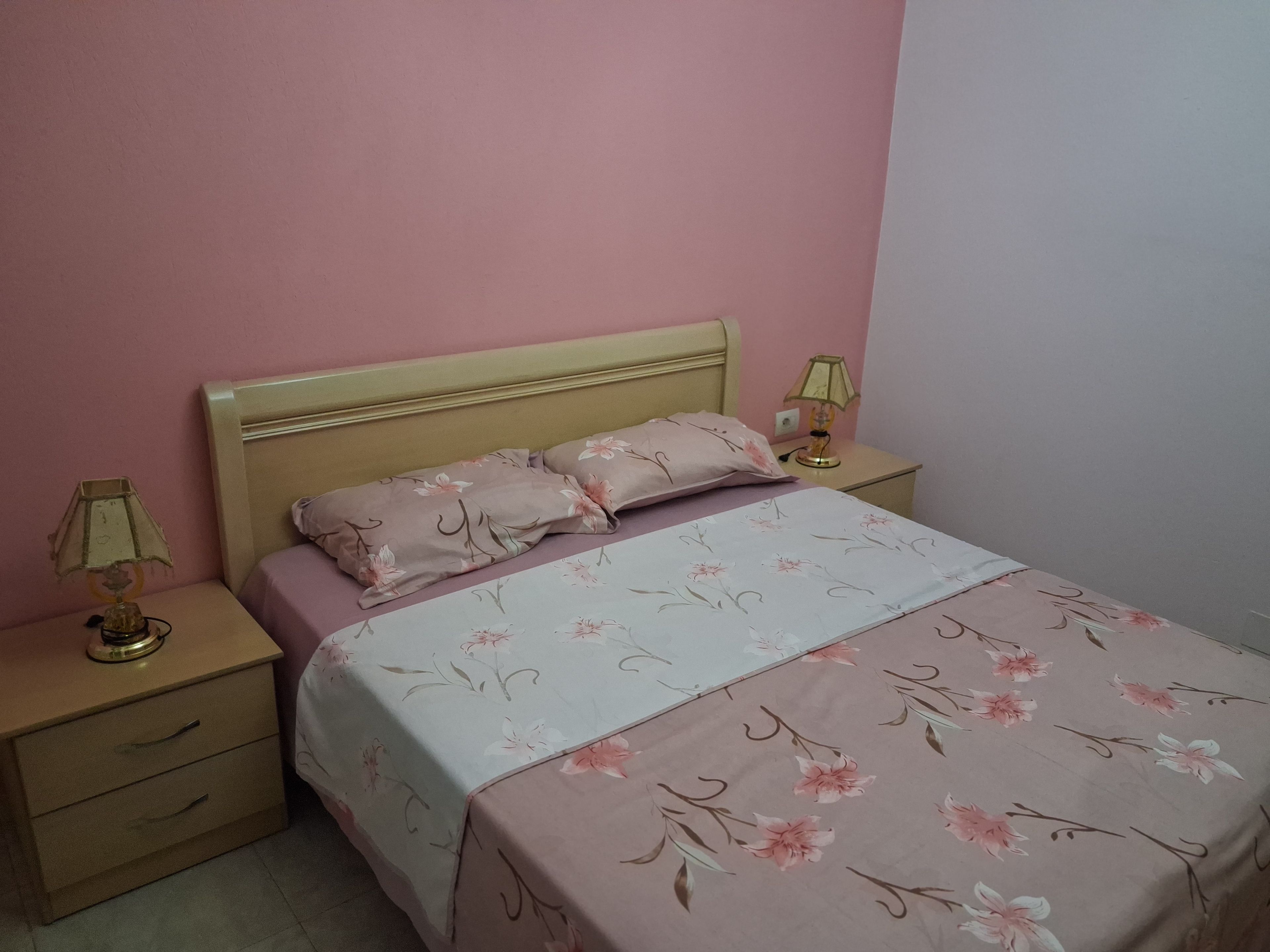 Ein Einzelbett mit floralem Überwurf, zwei Nachttischlampen, ein Nachttisch mit Schublade und ein Kopfteil an einer rosafarbenen Wand.