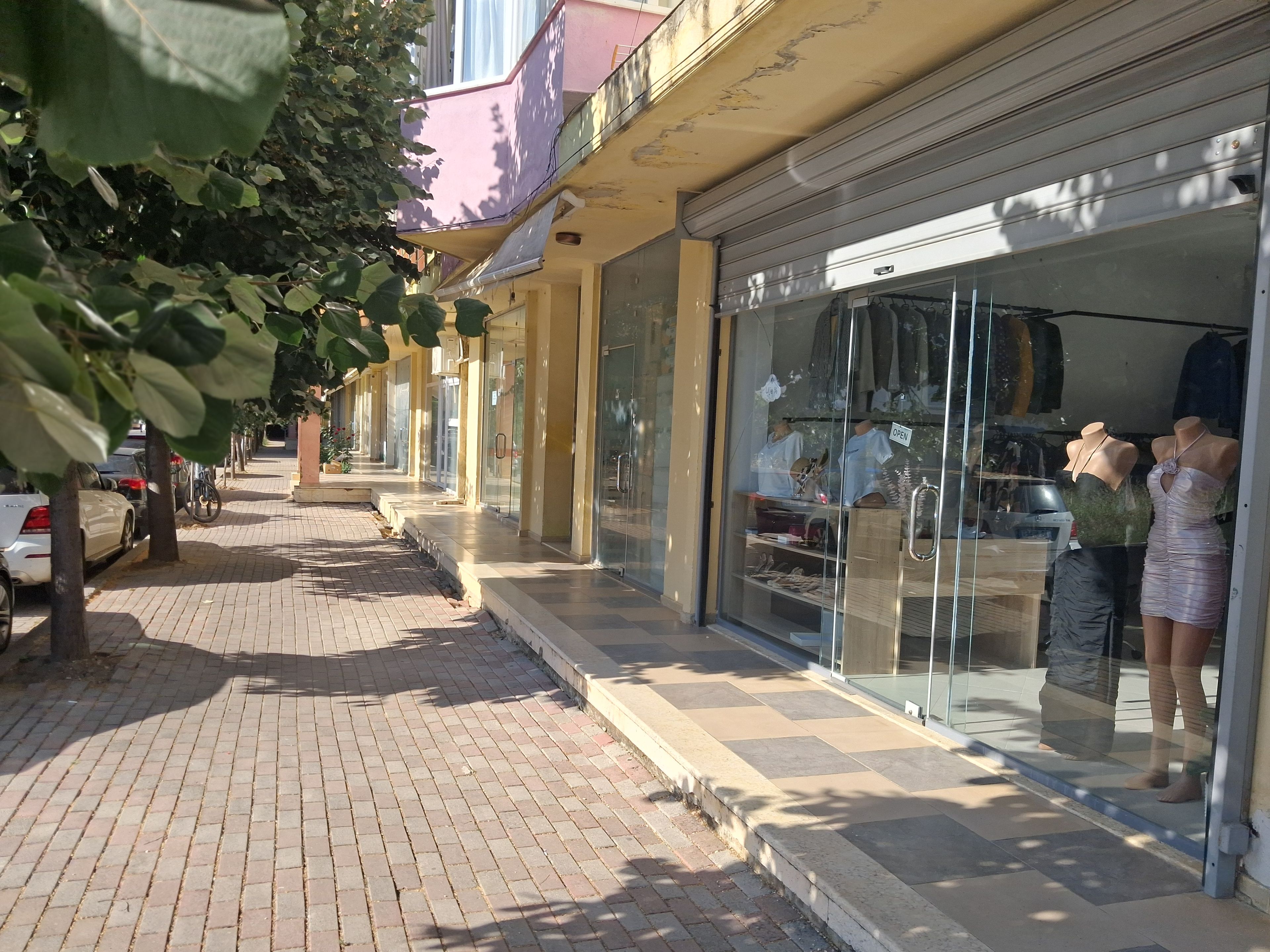 A storefront with mannequins displaying clothing, a paved sidewalk, and parked cars.
