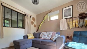 A living room with a grey sectional sofa, a blue armchair, a small wooden chair with a teddy bear, a woven rug, and a wall-mounted branch decoration.