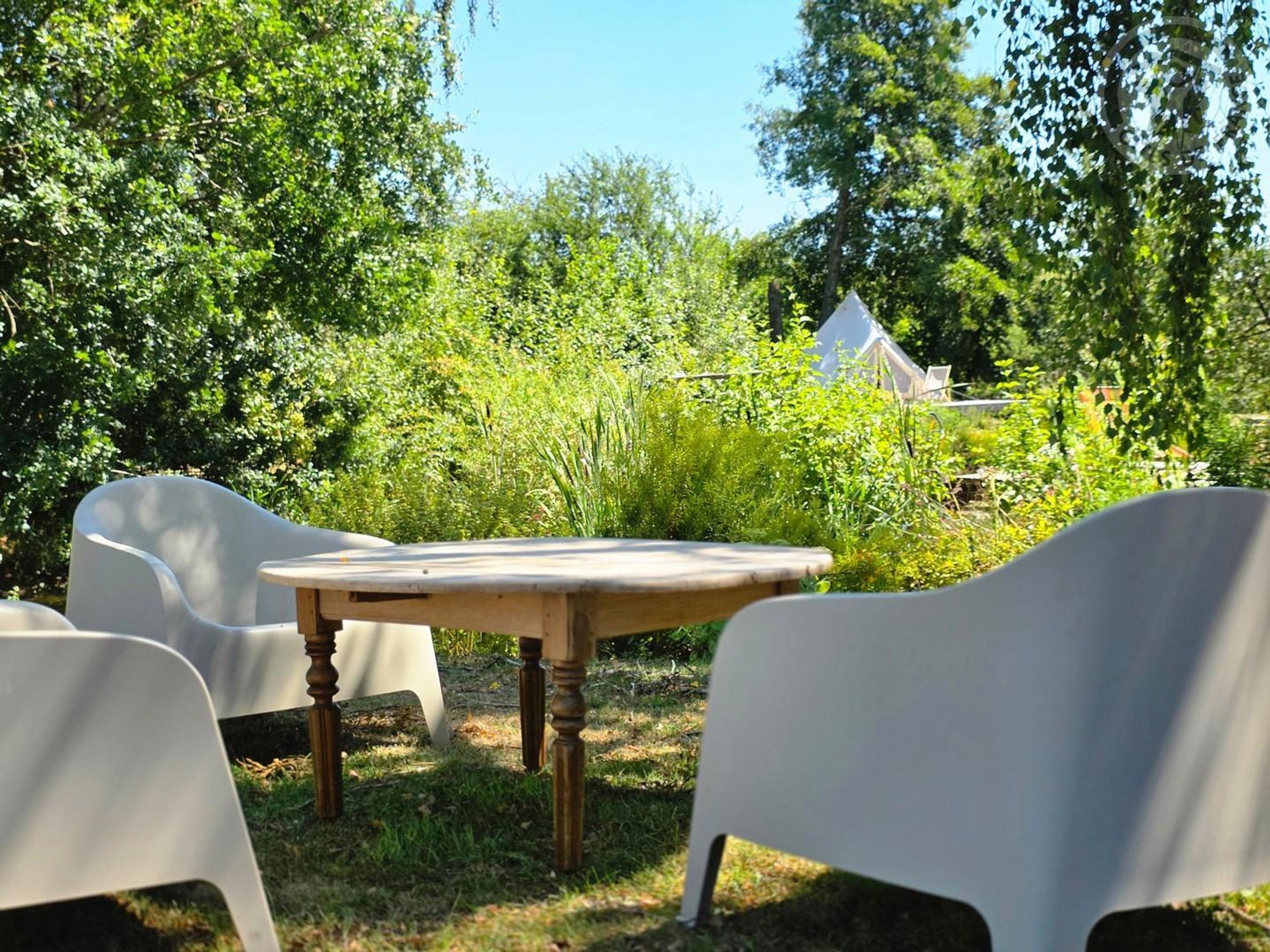 Un espace extérieur aménagé avec une table en bois et des chaises blanches, entouré d’une végétation luxuriante et agrémenté d’une tente en arrière-plan.