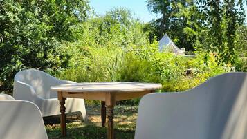 An outdoor seating area with a wooden table and white chairs, surrounded by lush greenery and a tent in the background.