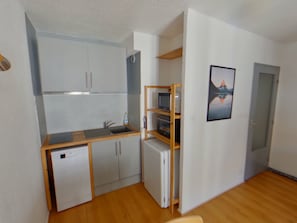 Eine kleine Küche mit Geschirrspüler, Spüle und kleinem Kühlschrank. Ein Holzbodenregal mit Fernseher und einem gerahmten Bild an der Wand.