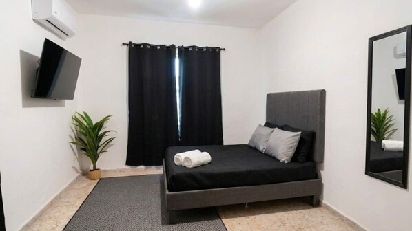 A modern bedroom with a bed, a flat-screen TV, a potted plant, and a window with curtains.