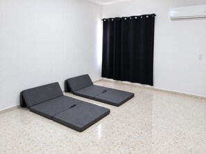 Ein Zimmer mit zwei dunkelgrauen, flachen Matratzen auf einem punktierten Boden, einem schwarzen Vorhang und einer weißen Wand mit einer an der Decke befestigten Klimaanlage.