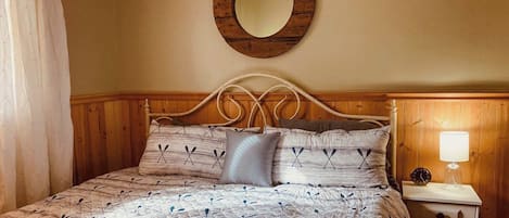 Une chambre à coucher avec une tête de lit en bois, un lit recouvert d’une literie à motifs, un miroir rond accroché au mur et une table de chevet avec une lampe.
