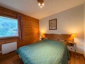A bedroom with a wooden headboard, a green quilted bedspread, a window with curtains, a radiator, and a wall-mounted picture. - Comfortable apartment with open view - 4 guests (Les Menuires)