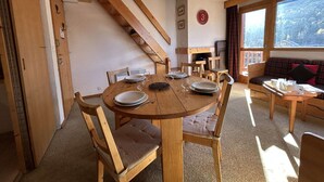 A cozy dining area with a wooden table set for four, surrounded by chairs with cushioned seats. A staircase with wooden railings is visible in the background, leading to an upper level. - Cozy apartment, pets welcome, near slopes - 5 guests (Méribel)