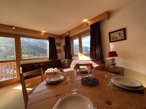 Ein Essbereich mit Holztisch, gedeckt für zwei Personen, einer Couch und Blick auf eine Berglandschaft durch große Fenster.