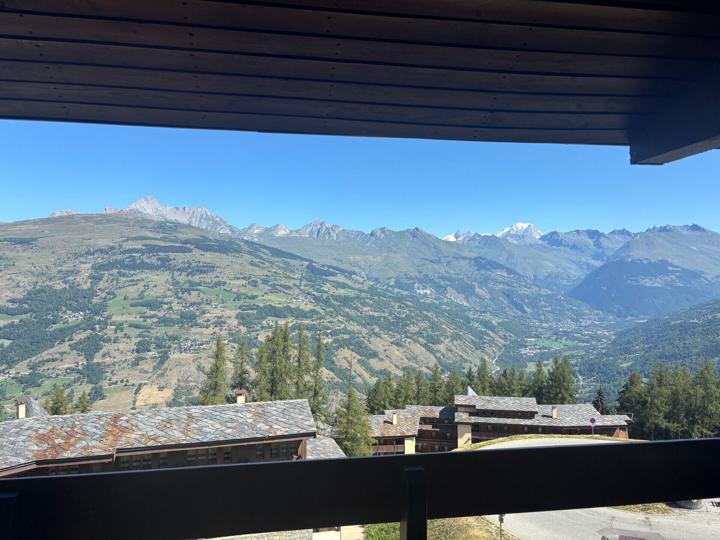 Une vue sur la montagne depuis un balcon doté d’une balustrade en bois, donnant sur une vallée avec des maisons et une chaîne de montagnes.