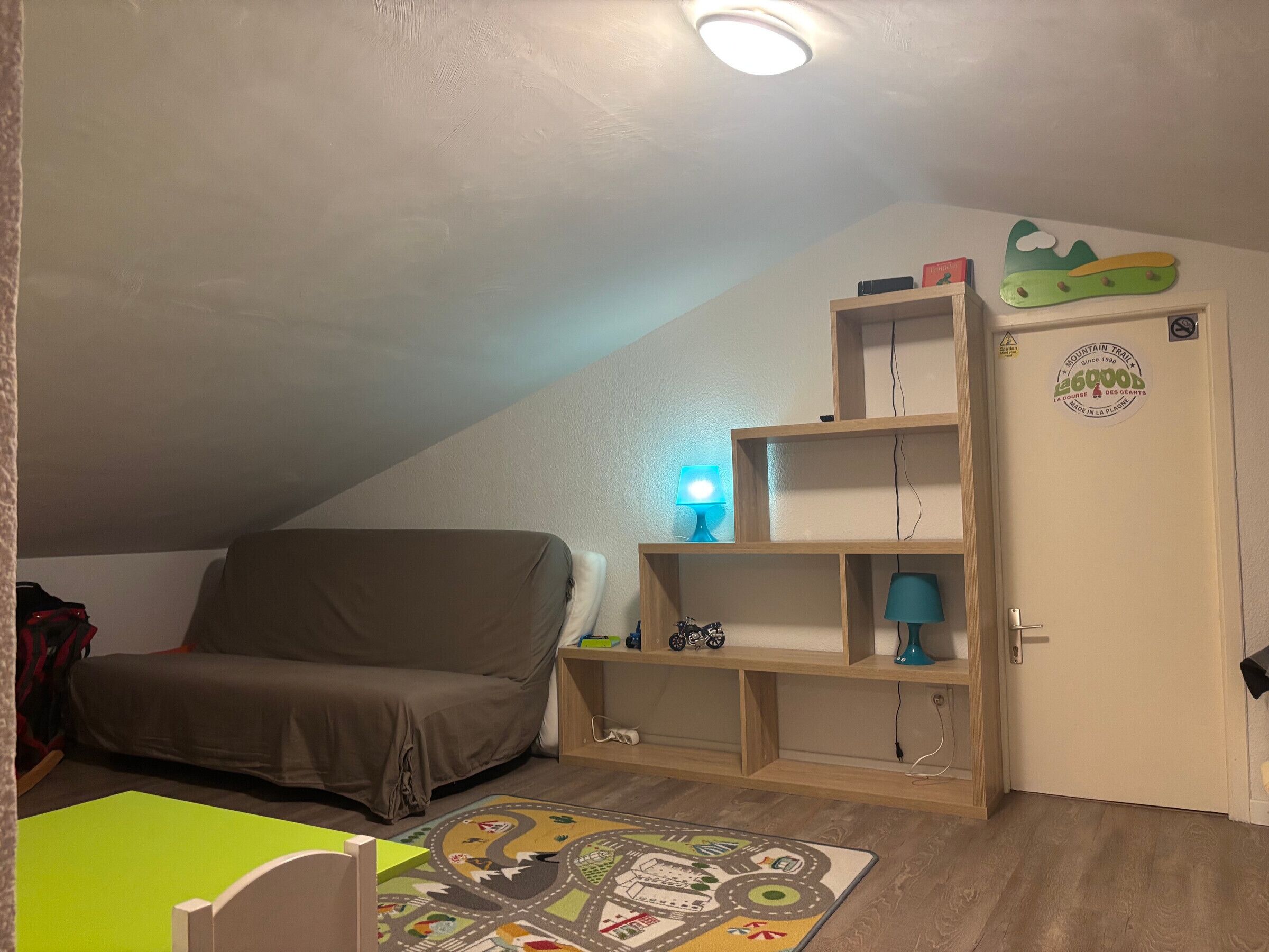 A room with a sloped ceiling, a sofa, a table, a lamp, a shelf with items, and a door with a sticker.