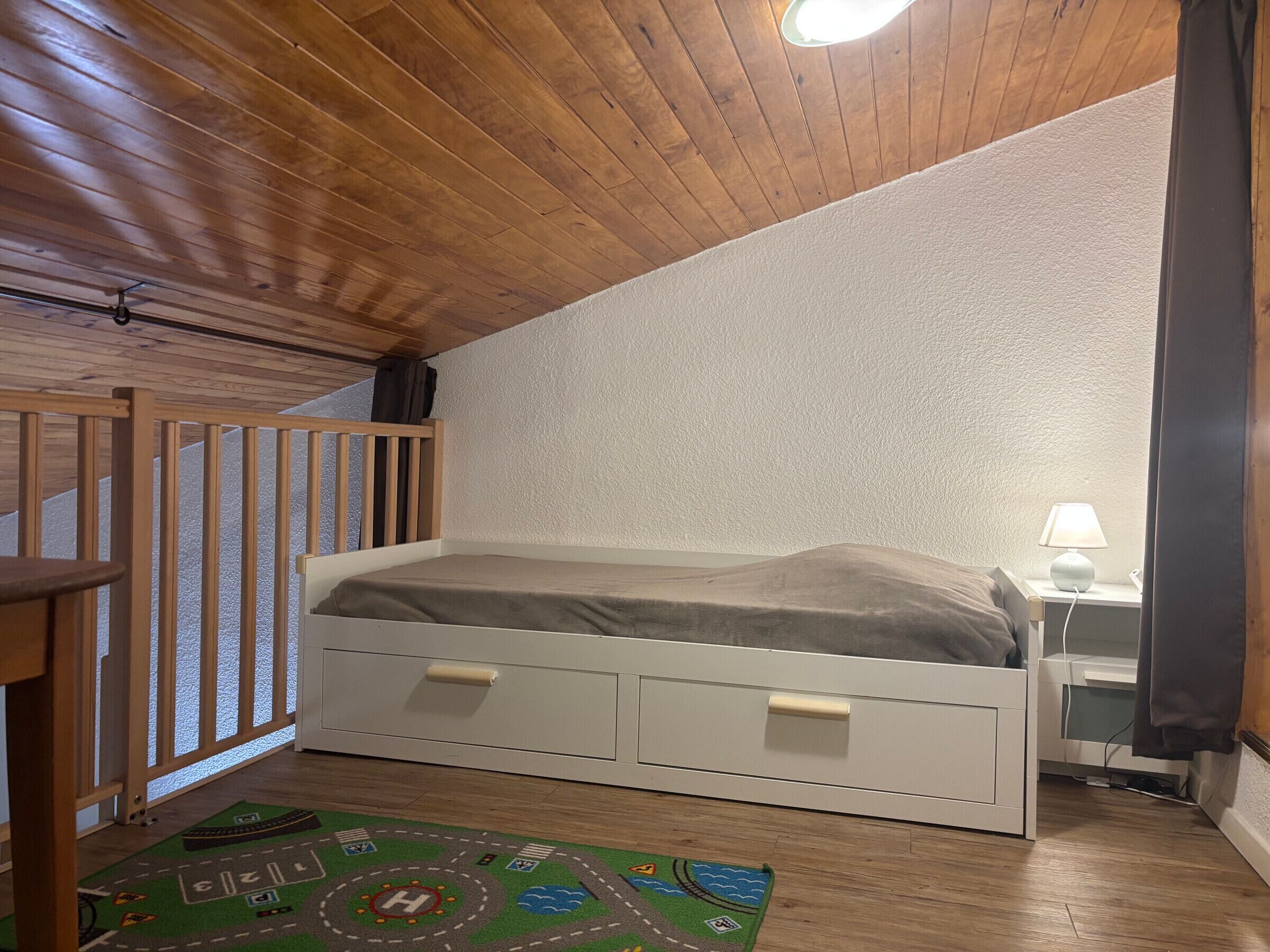 A bedroom with a wooden ceiling, a single bed with storage drawers, a nightstand with a lamp, and a colorful rug.