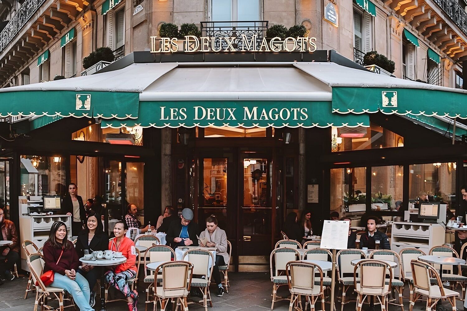 An outdoor café with tables and chairs, a sign reading 'LES DEUX MAGOTS,' and people dining.