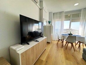 Ein modernes Wohnzimmer mit einem Flachbildfernseher, einem Essbereich mit Tisch und Stühlen und Blick auf die Stadt durch große Fenster.