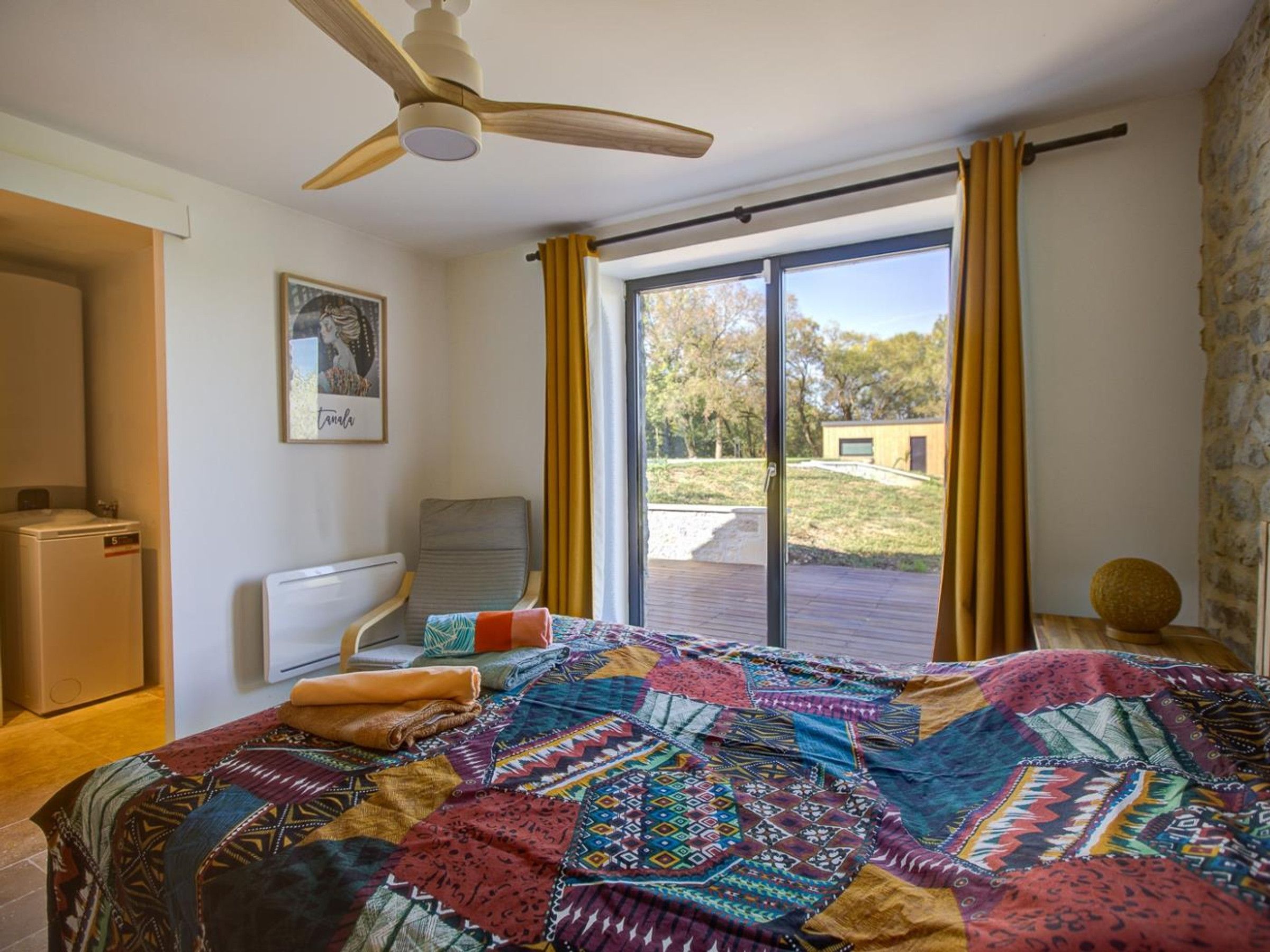 Ein Zimmer mit einem Bett, einem Sessel, einem Heizkörper, einem Deckenventilator, einem Fenster mit Vorhängen und Blick ins Freie.