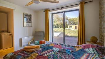 Ein Zimmer mit einem Bett, einem Sessel, einem Heizkörper, einem Deckenventilator, einem Fenster mit Vorhängen und Blick ins Freie.