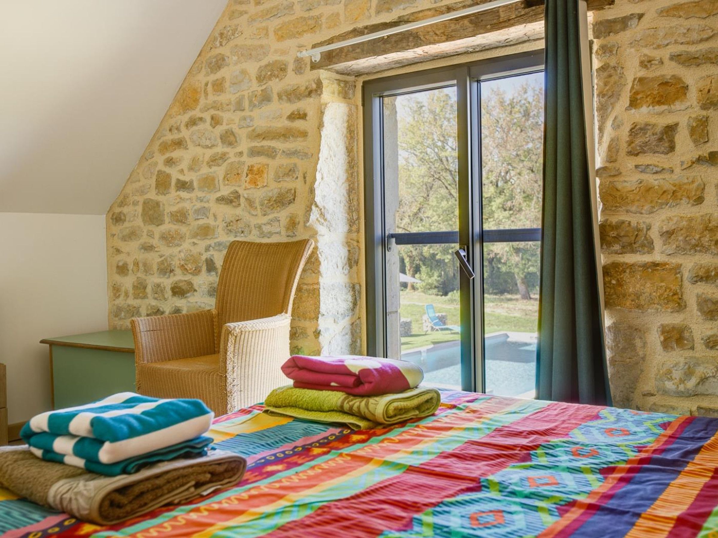 A room with a stone wall, a wicker chair, a bed with a colorful quilt, and a view of a swimming pool through a glass door.