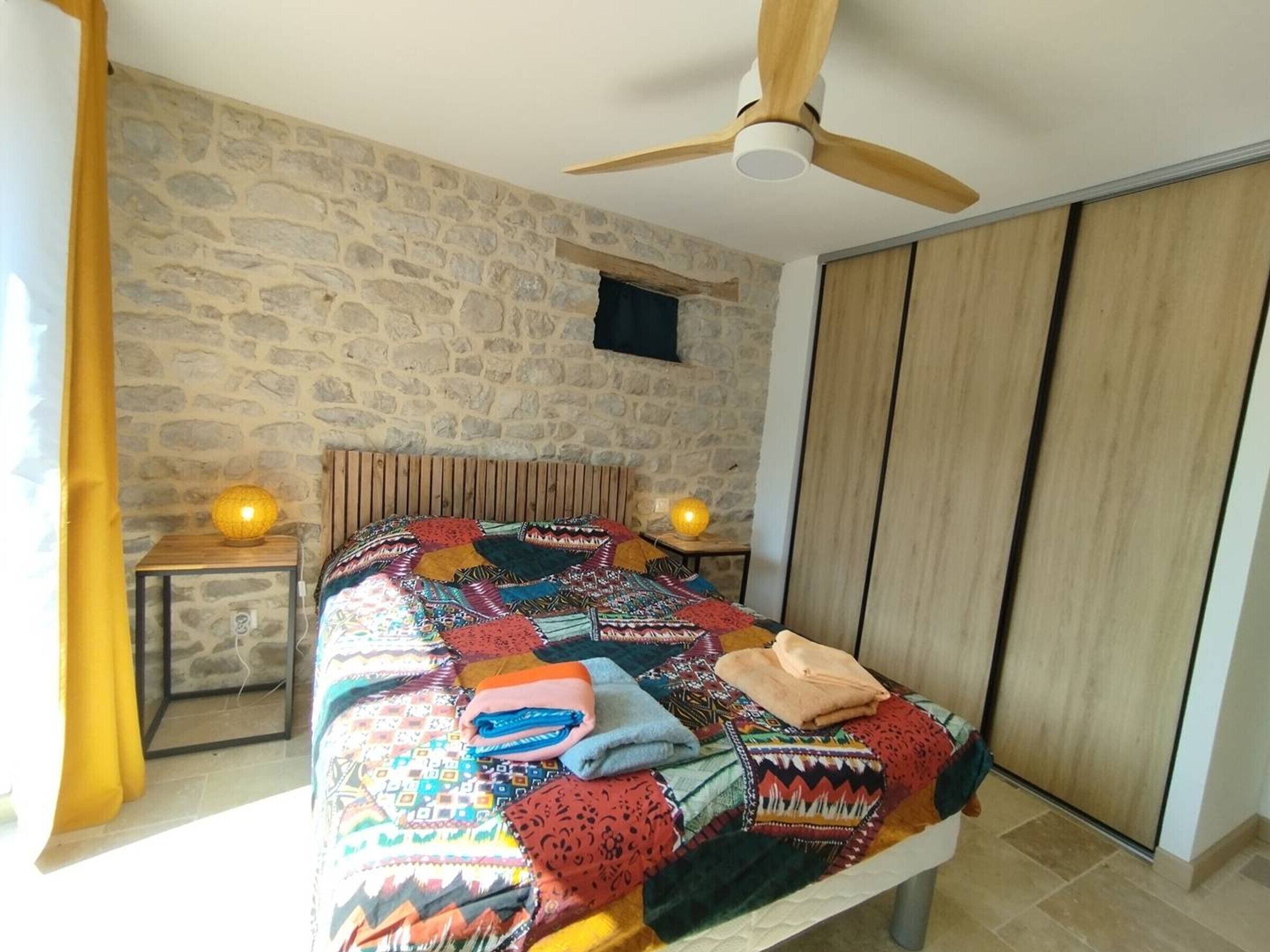 A bedroom with a stone wall, a wooden headboard, a bed with a colorful quilt, two bedside lamps, and a ceiling fan.
