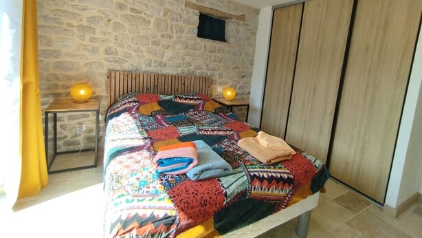 Quarto com parede de pedra, cabeceira de madeira, cama com colcha colorida, duas luminárias de cabeceira e um ventilador de teto.