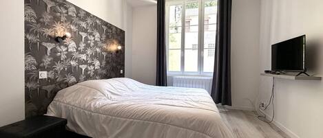 Ein Schlafzimmer mit einem Bett, einem am Wand montierten Fernseher, einem Fenster mit Vorhängen und einer Wandtapete mit Muster.