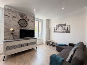 Ein modernes Wohnzimmer mit einem Flachbildfernseher auf einem weißen Schrank, einer Uhr an der Wand und einem großen Fenster.