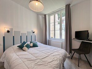 Ein Schlafzimmer mit Bett, Fernseher auf einem Schrank, einem Fenster mit Vorhängen und einer Pendelleuchte.