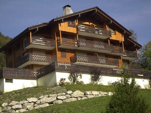 Un chalet à plusieurs étages, doté de balcons, situé sur une colline et adossé à un mur de soutènement en pierre.
